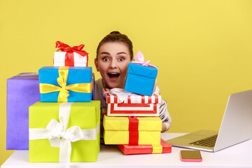 Extremely excited woman sitting at workplace among present boxes, looking at camera with open mouth, being surprised with many gifts. Indoor studio studio shot isolated on yellow background.