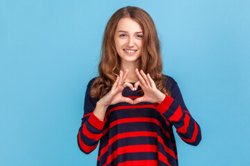 Romantic woman wearing striped casual style sweater making heart shape with fingers, gesturing love hope charity sign, looks at camera. Indoor studio shot isolated on blue background.
