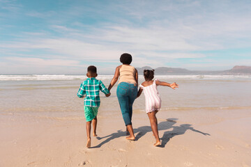 Fototapeta na wymiar Rear view of african american grandmother holding boy and girl's hands running at beach against sky