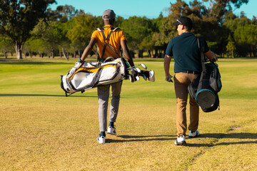 Rear view of multiracial young male friends with golf bags walking on land at golf course in summer