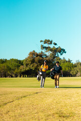Multiracial young male friends with golf bags walking at golf course against clear sky in summer