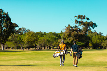 Rear view of multiracial young male friends with golf bags walking at golf course against clear sky - Powered by Adobe