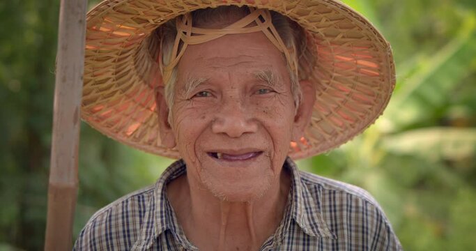 A happy smiling Asian male farmer who is a white-haired elderly person wearing a traditional woven hat made of leaves holding a bamboo-handled spade stands in vegetable plantation at countryside.