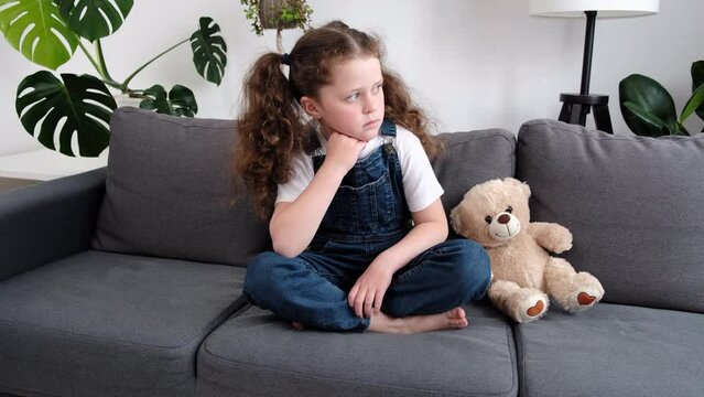 Sad lonely kid sitting alone on grey couch in living room with teddy bear toy, feeling depressed, upset child looking at side, going through trauma. Childhood problems, abuse, in family concept