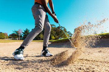 Low section of african american young man hitting golf ball with club on sand against clear sky - Powered by Adobe