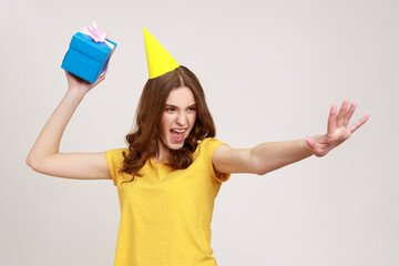 Catch your present! Take it! Happy carefree young female in yellow t-shirt ready to throw birthday box, having fun and sharing gift. Indoor studio shot isolated on gray background.