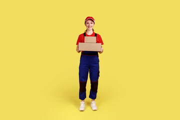 Fototapeta na wymiar Full length portrait of courier woman standing with two parcels in hands, looking at camera, delivery service, wearing overalls and red cap. Indoor studio shot isolated on yellow background.