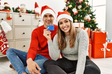 Young couple smiling happy wearing christmas hat. Sitting on the floor holding credit card at home.