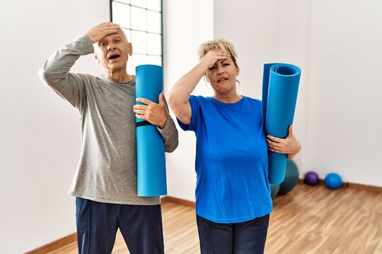 Middle age caucasian couple holding yoga mat at pilates room stressed and frustrated with hand on head, surprised and angry face
