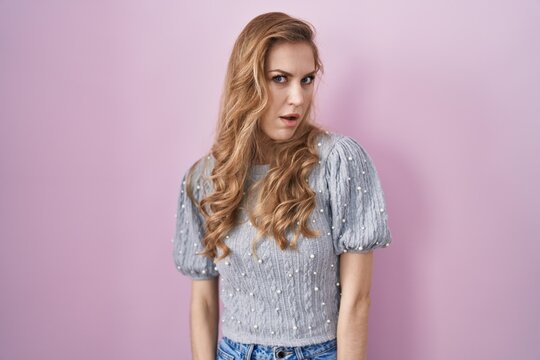 Beautiful blonde woman standing over pink background in shock face, looking skeptical and sarcastic, surprised with open mouth