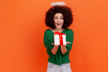 Excited woman with Afro hairstyle wearing green casual style sweater and nimb over head holding out present, giving gift, positive expression. Indoor studio shot isolated on orange background.