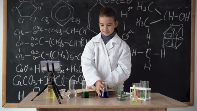 ittle scientist girl in white coat leads an online video blog, records video using smartphone. Research and education. Chemical laboratory with glass flasks. Schoolgirl conducts a chemical experiment