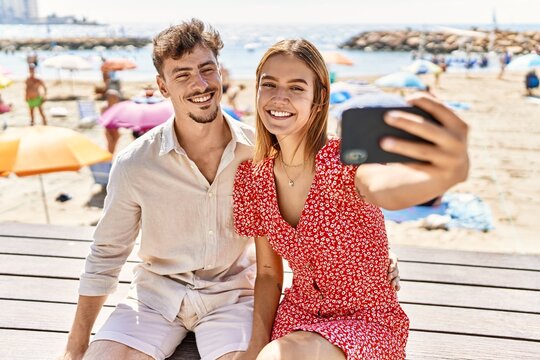 Young hispanic couple on vacation make selfie by the smartphone sitting on bench at the beach