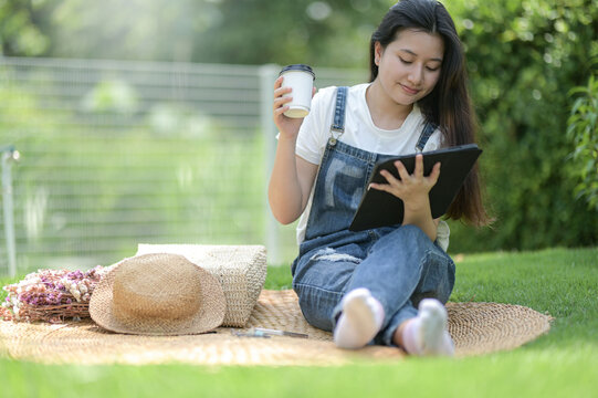 A teenage woman sits in the grass relaxing with a tablet and a cup of coffee, Chatting online.