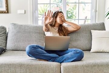 Beautiful young brunette woman sitting on the sofa using computer laptop at home covering eyes with...