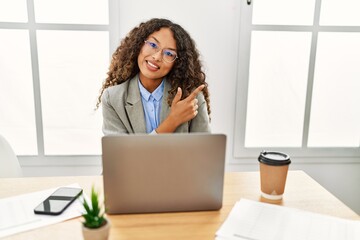Beautiful hispanic business woman sitting on desk at office working with laptop with a big smile on face, pointing with hand and finger to the side looking at the camera.