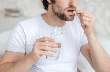 Sad young caucasian man with stubble wake up on white bed, holds glass of water and take pill in...