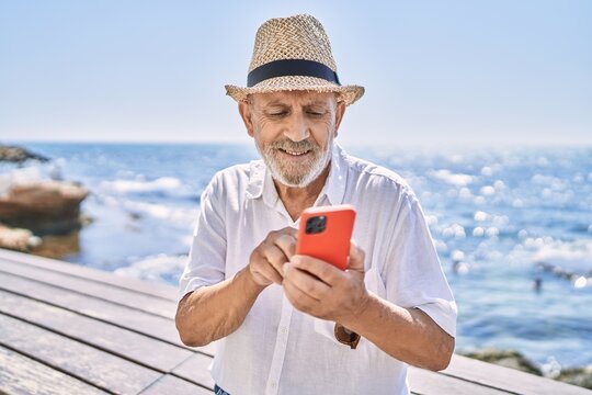 Senior man smiling confident wearing summer hat using smartphone at seaside