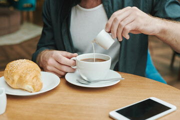 Busy young bearded man sitting at table in modern cafe and drinking coffee while discussing project by phone