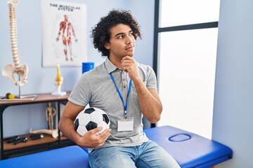 Hispanic man with curly hair working as football physiotherapist with hand on chin thinking about...