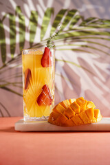 tall cocktail glass filled with yellow juice and fresh strawberries and cut in cubes mango half on tropical palm tree background, gobo mask effect