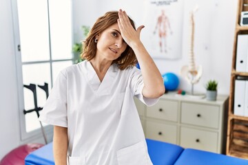 Middle age physiotherapist woman working at pain recovery clinic surprised with hand on head for...