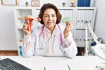 Middle age hispanic doctor woman holding anatomical female genital organ and birth control pills...