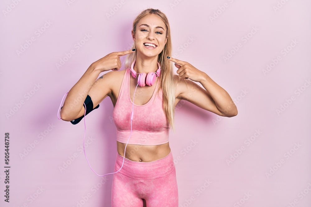 Sticker Young beautiful caucasian woman wearing gym clothes and using headphones smiling cheerful showing and pointing with fingers teeth and mouth. dental health concept.