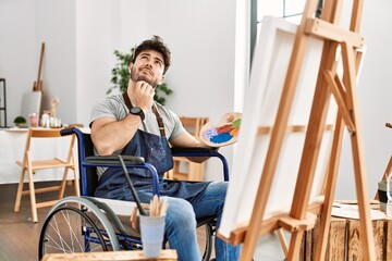 Young hispanic man sitting on wheelchair painting at art studio thinking concentrated about doubt...