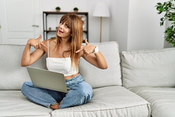 Hispanic woman sitting on the sofa at home using laptop looking confident with smile on face,...