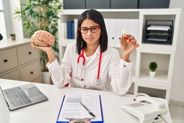 Young hispanic doctor woman holding brain and cbd oil skeptic and nervous, frowning upset because...