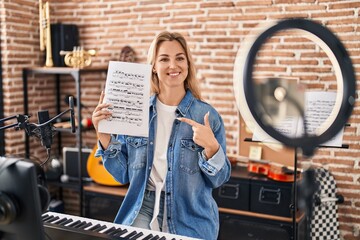 Young caucasian woman doing online music tutorial showing music sheet smiling happy pointing with...