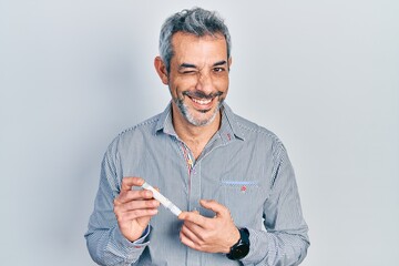 Handsome middle age man with grey hair holding glucometer device winking looking at the camera with...