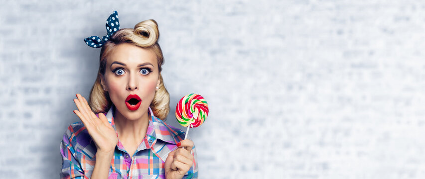 Excited surprised woman with lollipop. Girl pin up with open mouth. Blond model at retro fashion and vintage concept. White brick wall background. Wide copy space for some slogan or text.