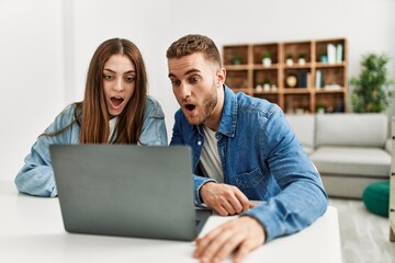 Young caucasian couple working using computer laptop at home scared and amazed with open mouth for surprise, disbelief face