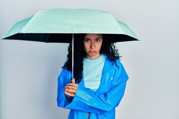 Young hispanic woman with curly hair wearing a raincoat and umbrella depressed and worry for...