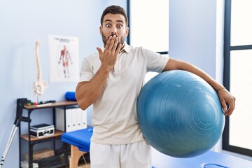 Handsome hispanic man holding pilates ball at rehabilitation clinic covering mouth with hand,...