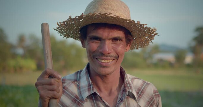 A happy smiling Asian farmer who is indigenous, wearing a straw hat, holding a hoe, mowing the grass and weeds in the newly planted corn plots during the evening light at the countryside.

