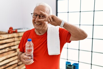 Senior man wearing sportswear and towel at the gym doing peace symbol with fingers over face,...