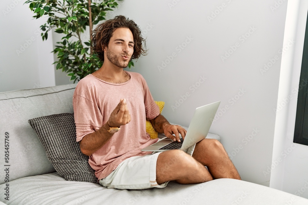 Sticker young hispanic man sitting on the sofa at home using laptop doing money gesture with hands, asking f