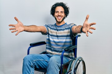 Handsome hispanic man sitting on wheelchair looking at the camera smiling with open arms for hug....