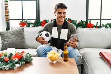 Young hispanic man supporting soccer game using smartphone sitting on sofa by christmas decor at...