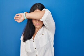 Young hispanic woman standing over blue background covering eyes with arm, looking serious and sad. sightless, hiding and rejection concept