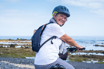 Happy cyclist senior woman along the sea beach carrying a backpack, wearing helmet running with...