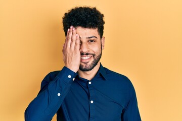 Young arab man with beard wearing casual shirt covering one eye with hand, confident smile on face and surprise emotion.