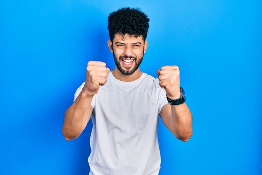 Young arab man with beard wearing casual white t shirt angry and mad raising fists frustrated and furious while shouting with anger. rage and aggressive concept.