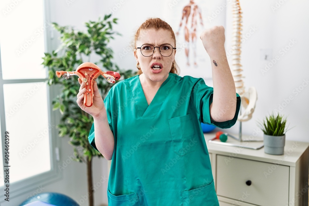 Poster Young redhead doctor woman holding anatomical model of female genital organ annoyed and frustrated shouting with anger, yelling crazy with anger and hand raised