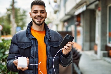 Handsome hispanic man with beard smiling happy and confident at the city wearing winter coat using smartphone battery charger