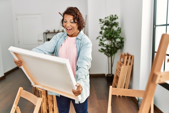 Middle age hispanic artist woman smiling happy holding canvas at art studio.