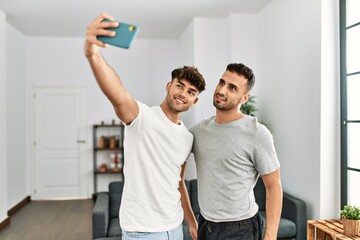 Two hispanic men couple making selfie by the smartphone hugging each other at home